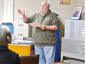 Photo of Steve Chappell with a green jumper with a hood. Holding some papers and his hand is forward