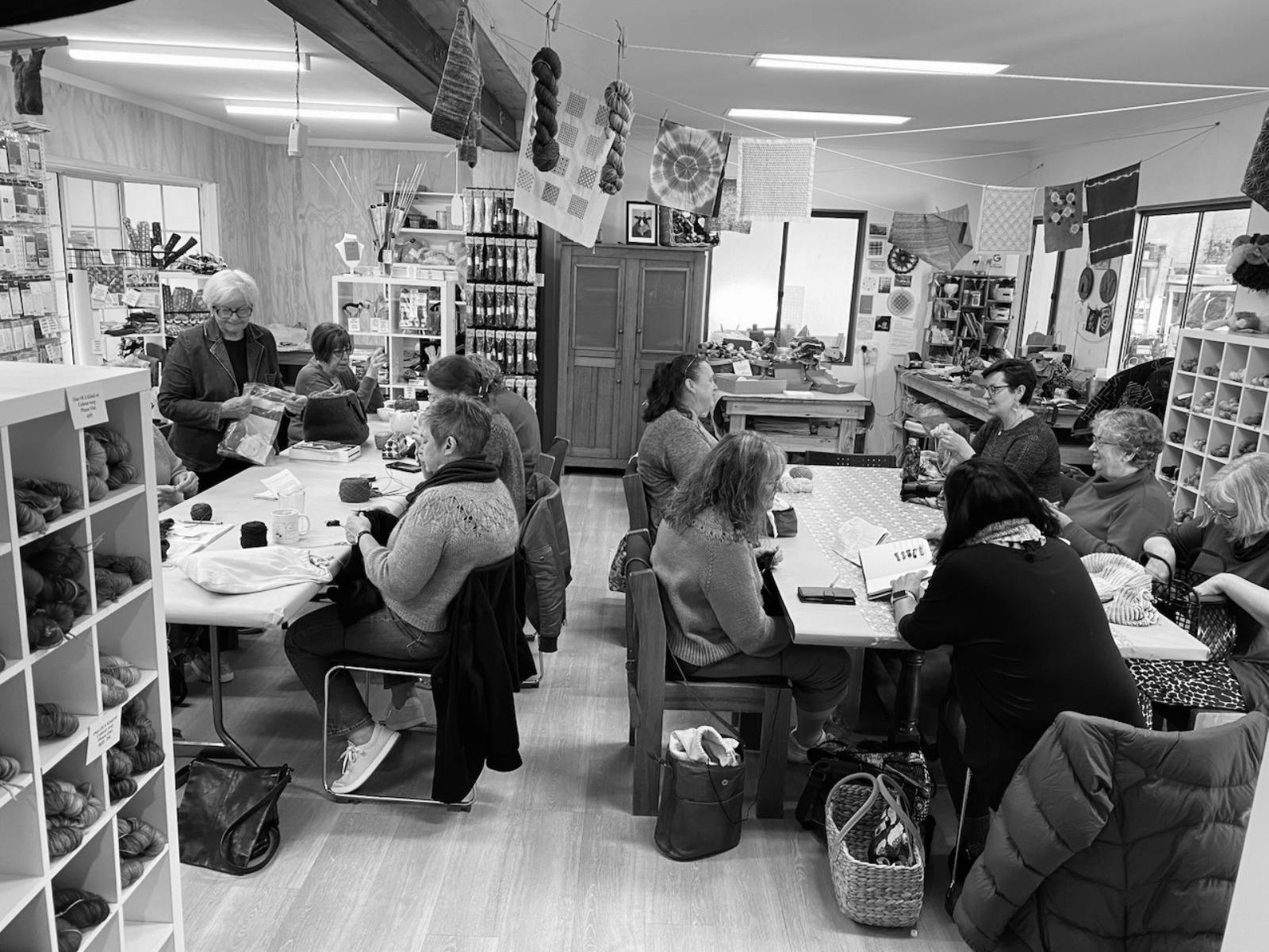 A black and white image of the Bombed Yarns studio, where people sit around two different tables.
