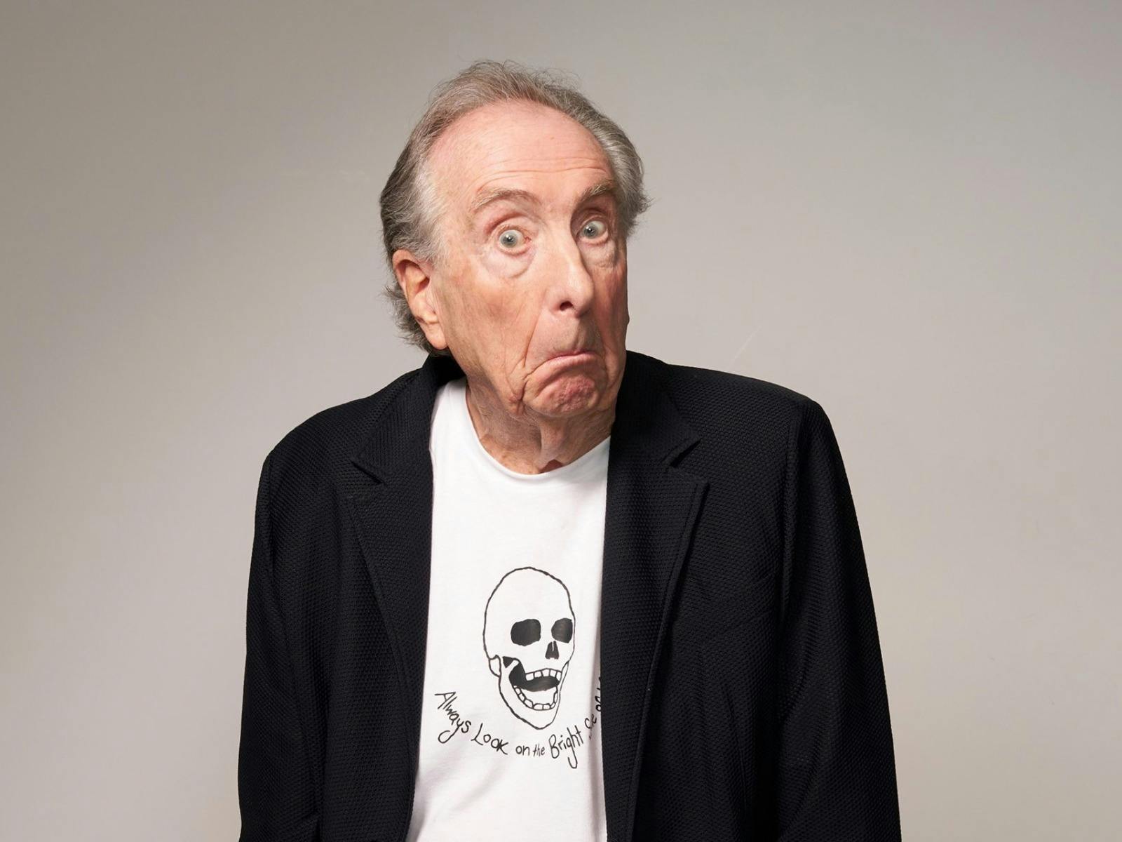 Eric Idle pulls a surprised, wide-eyed expression in a black blazer over a t-shirt