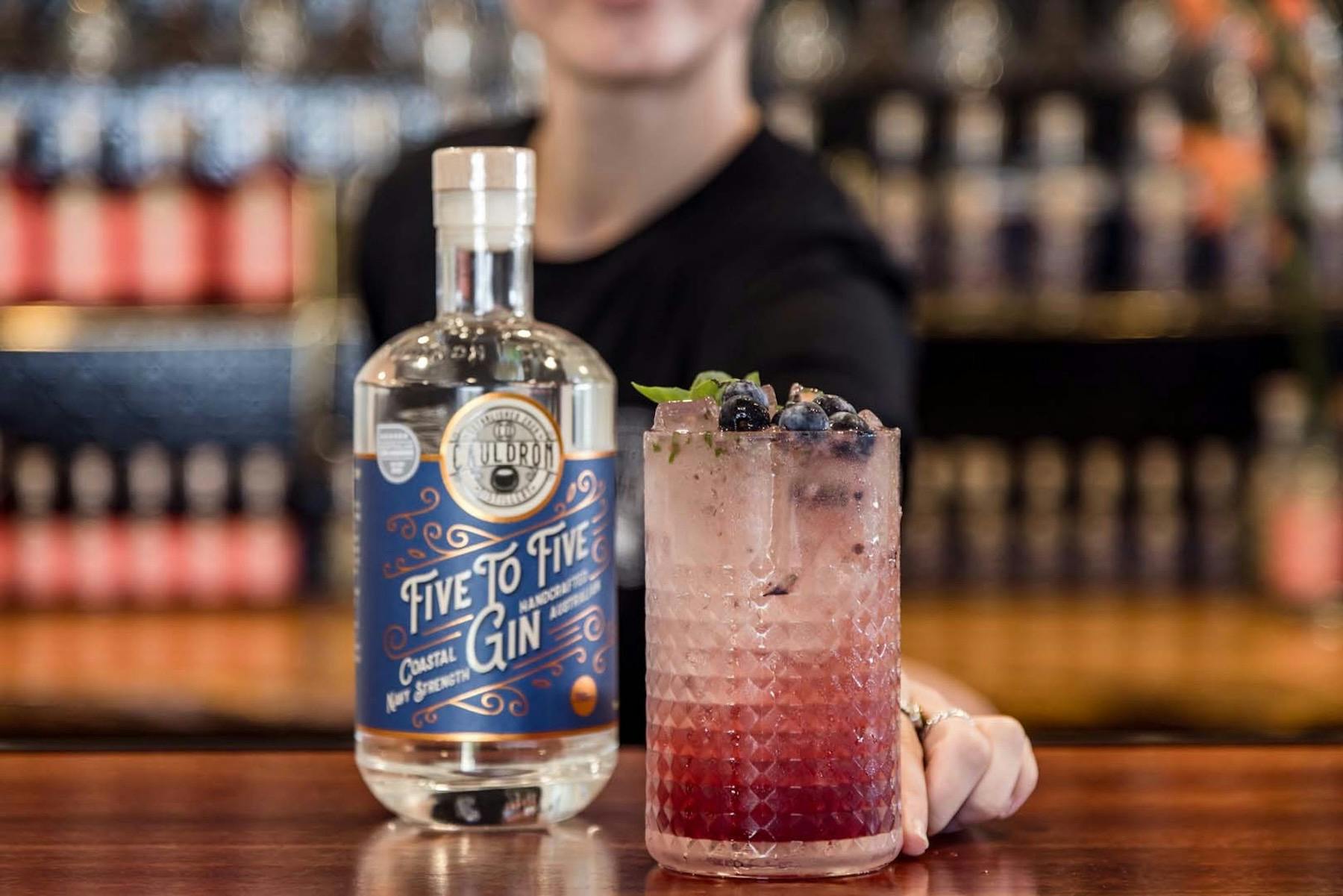 A bar maid holding a purple gin cocktail on the bar next to a bottle of gin