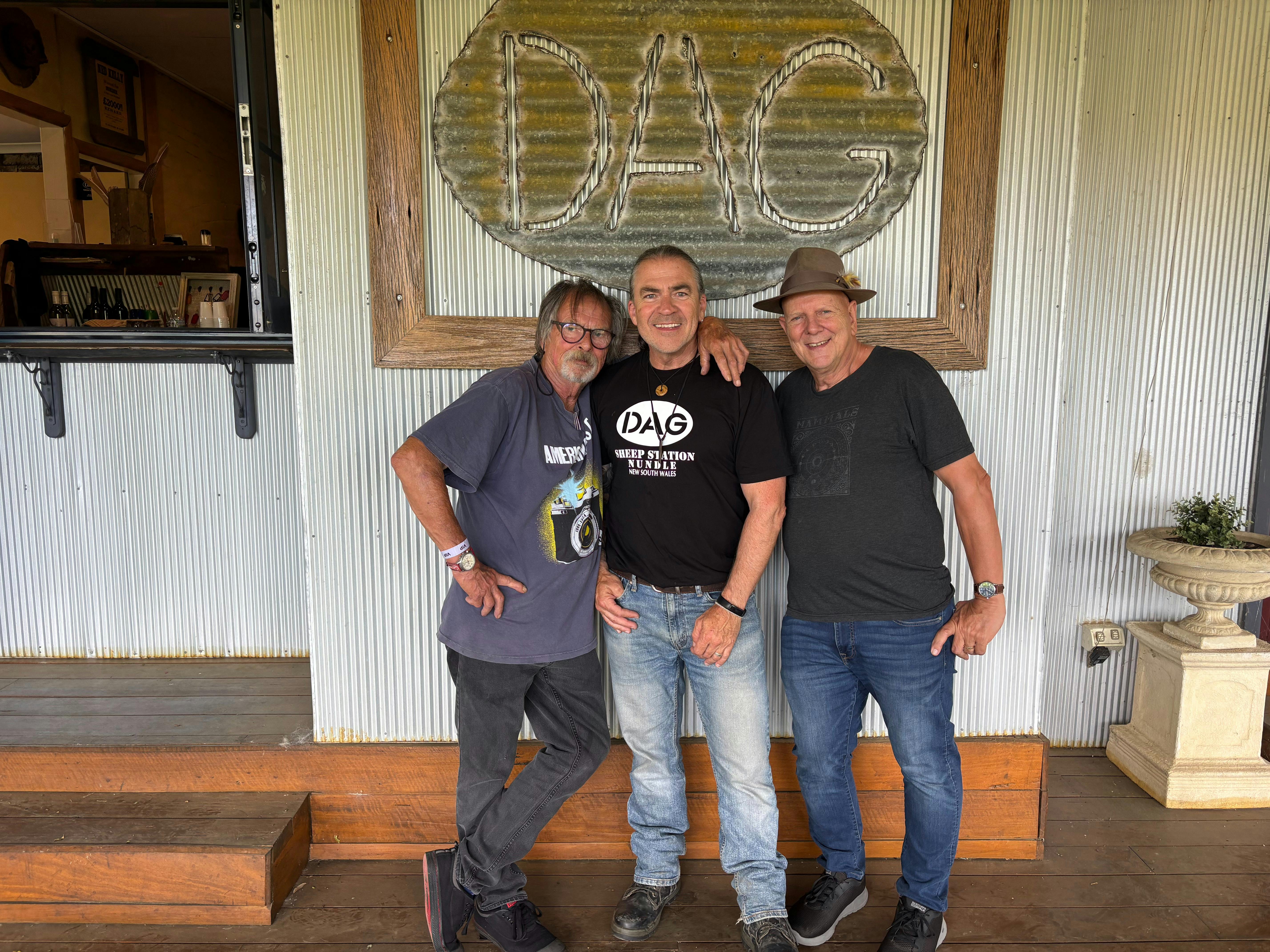 3 happy men standing in front of a sign which says "DAG" on a corrugated iron wall.