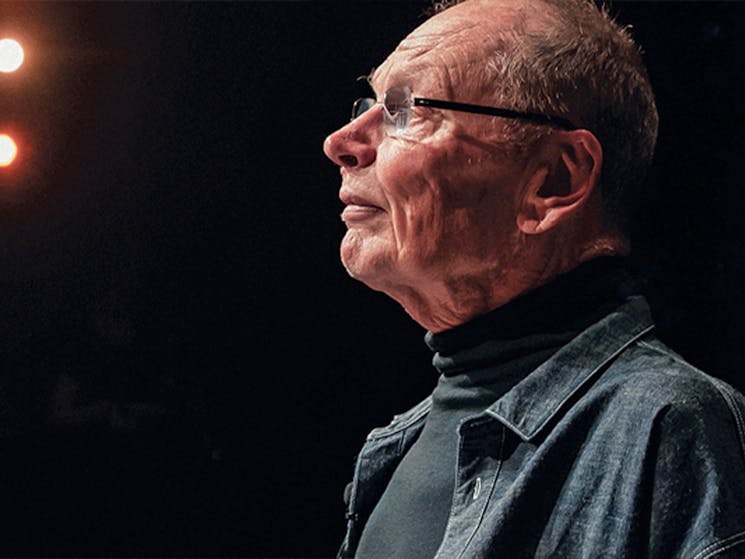 An older man in glasses, John Bell, looks pensively outwards. His face is light by stage lights.