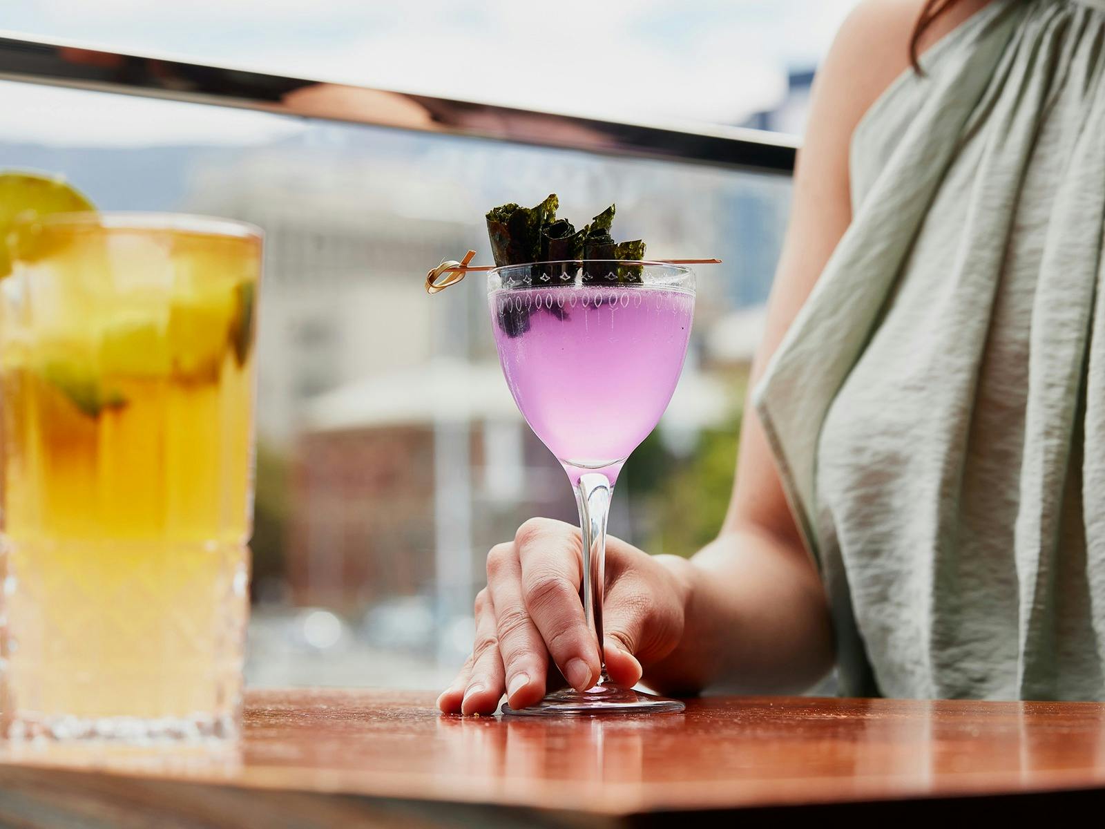 A yellow and purple cocktail on a table, with a lady's hand holding one.