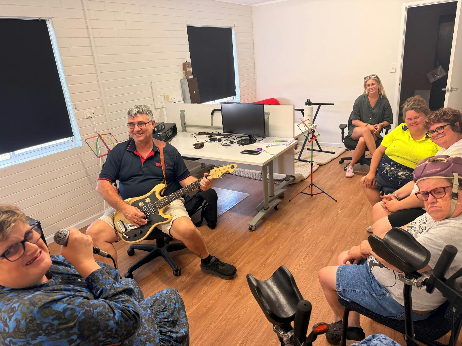 A small group participates in an indoor music session in an office-style room. One person plays an e