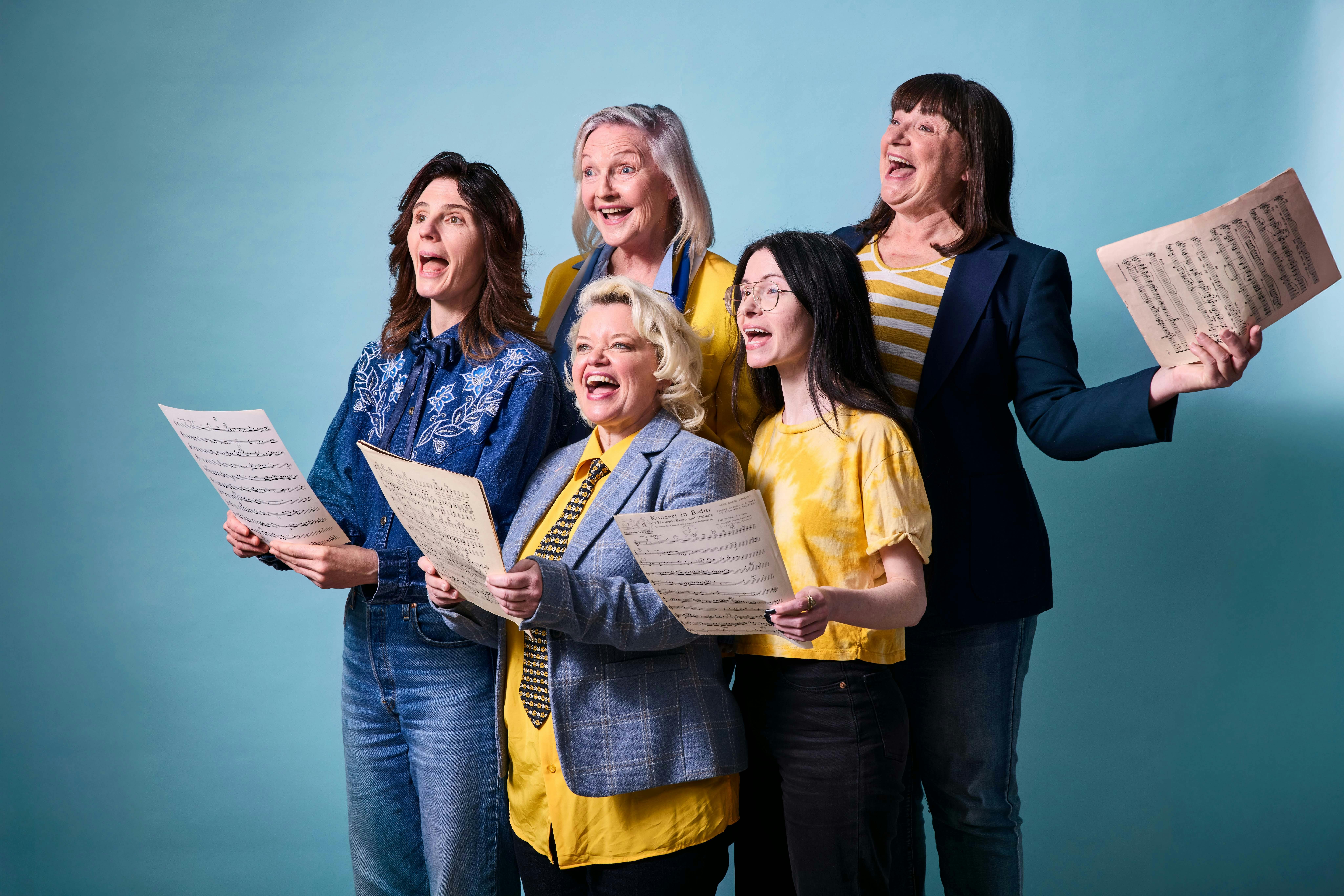 Five woman hold pieces of sheet music and sing happily