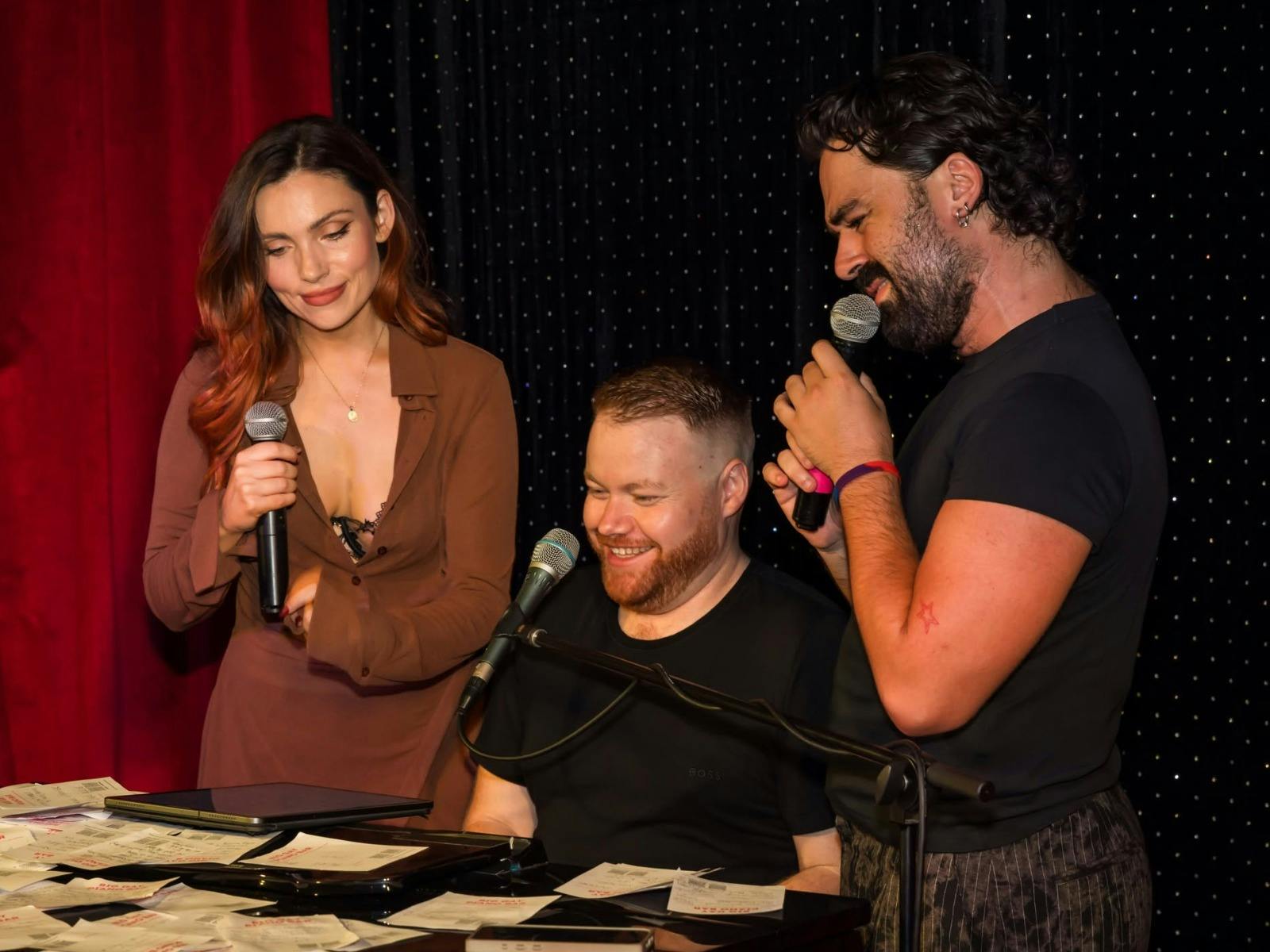 Two singers holding microphones beside a smiling pianist at a keyboard.