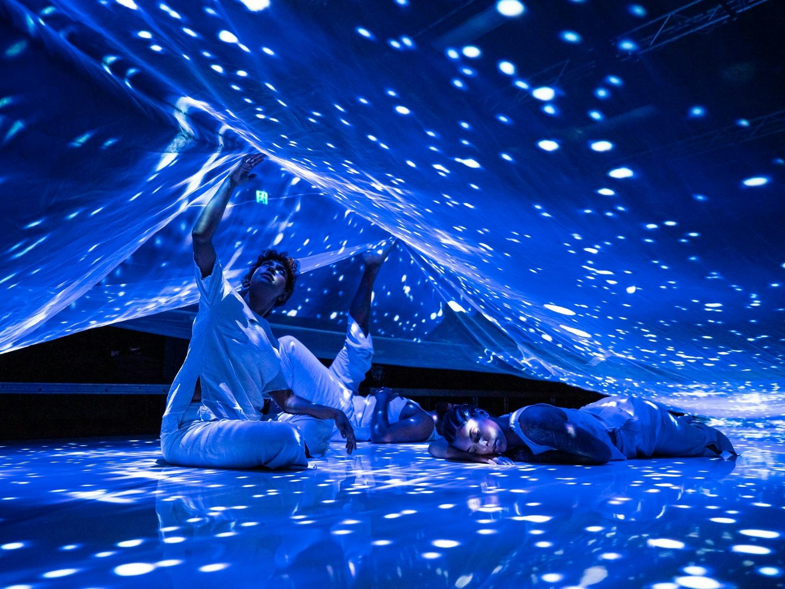 Three people relax beneath a sheet that is illuminated by many star-like spots and blue light.
