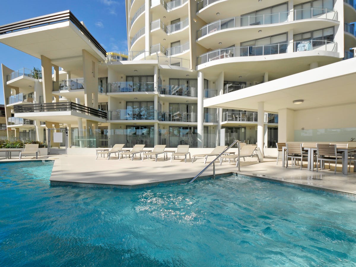 Sparkling blue pool with lounges and BBQ area, framed by the elegant building and clear blue sky.