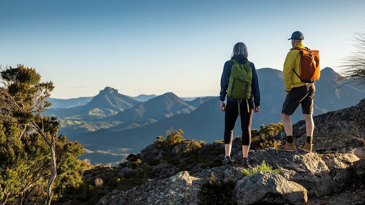 Two hikers stand on the top of a mountain, looking towards another mountain