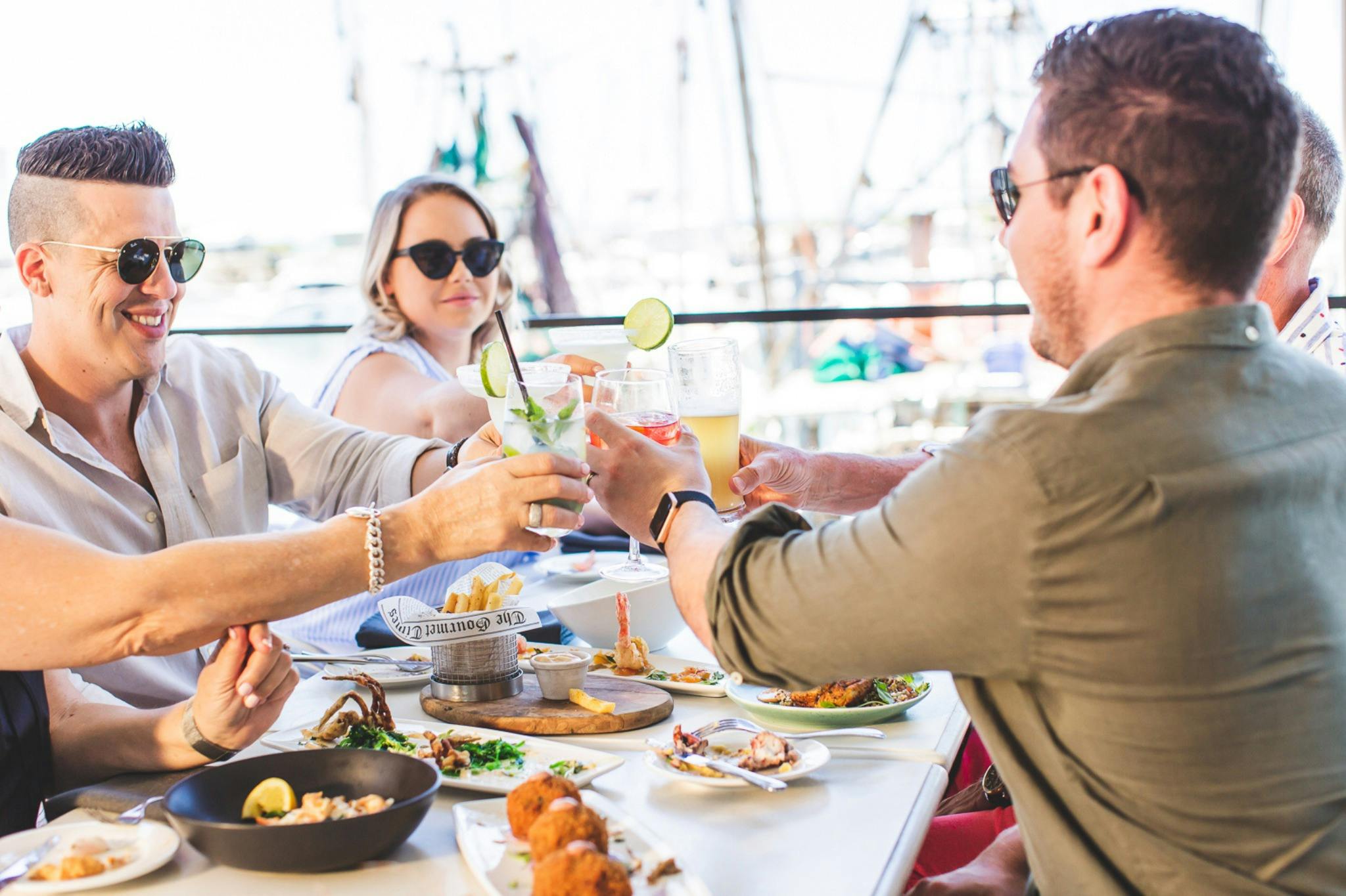 Friends clinking drinks around a table of food overlooking the harbour
