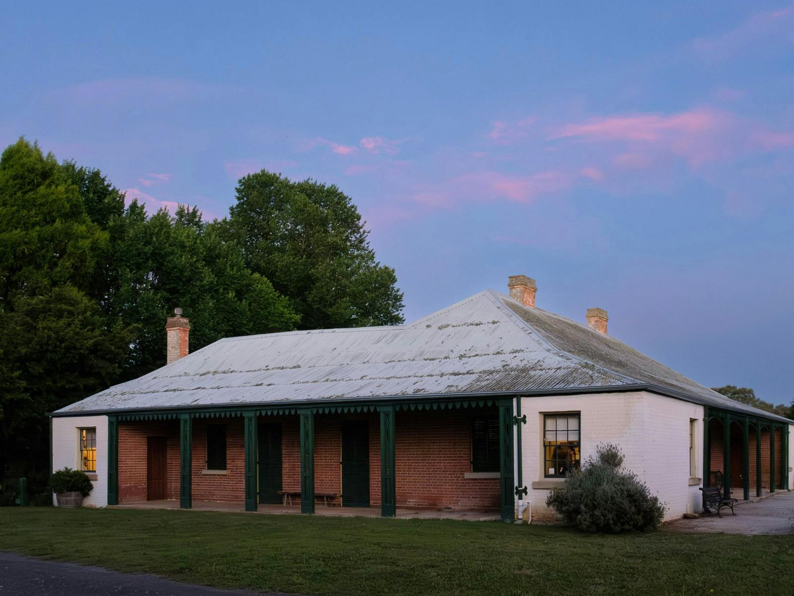 Pink clouds hang above a historic one-storey building. The building has a tin roof and red bricks.