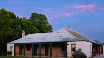 Pink clouds hang above a historic one-storey building. The building has a tin roof and red bricks.