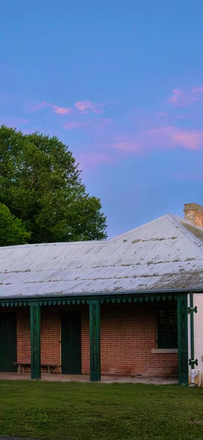 Pink clouds hang above a historic one-storey building. The building has a tin roof and red bricks.