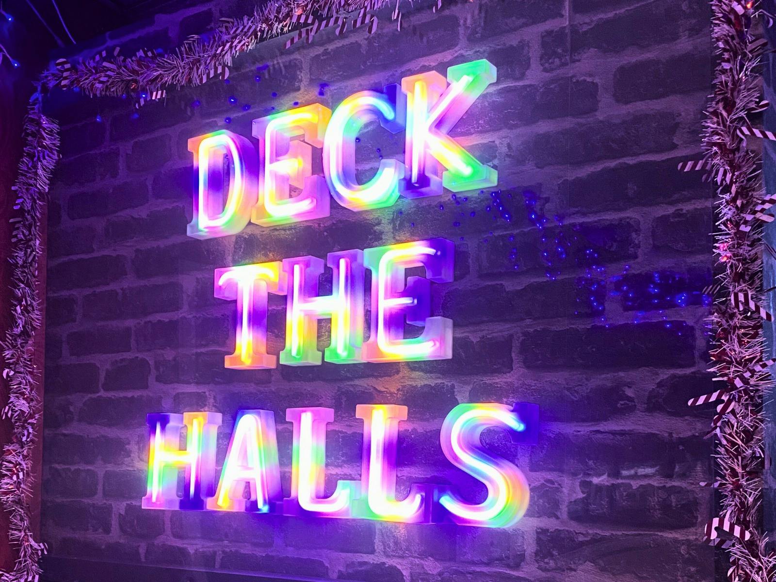Illuminated letters read "DECK THE HALLS" on a faux-brick wall
