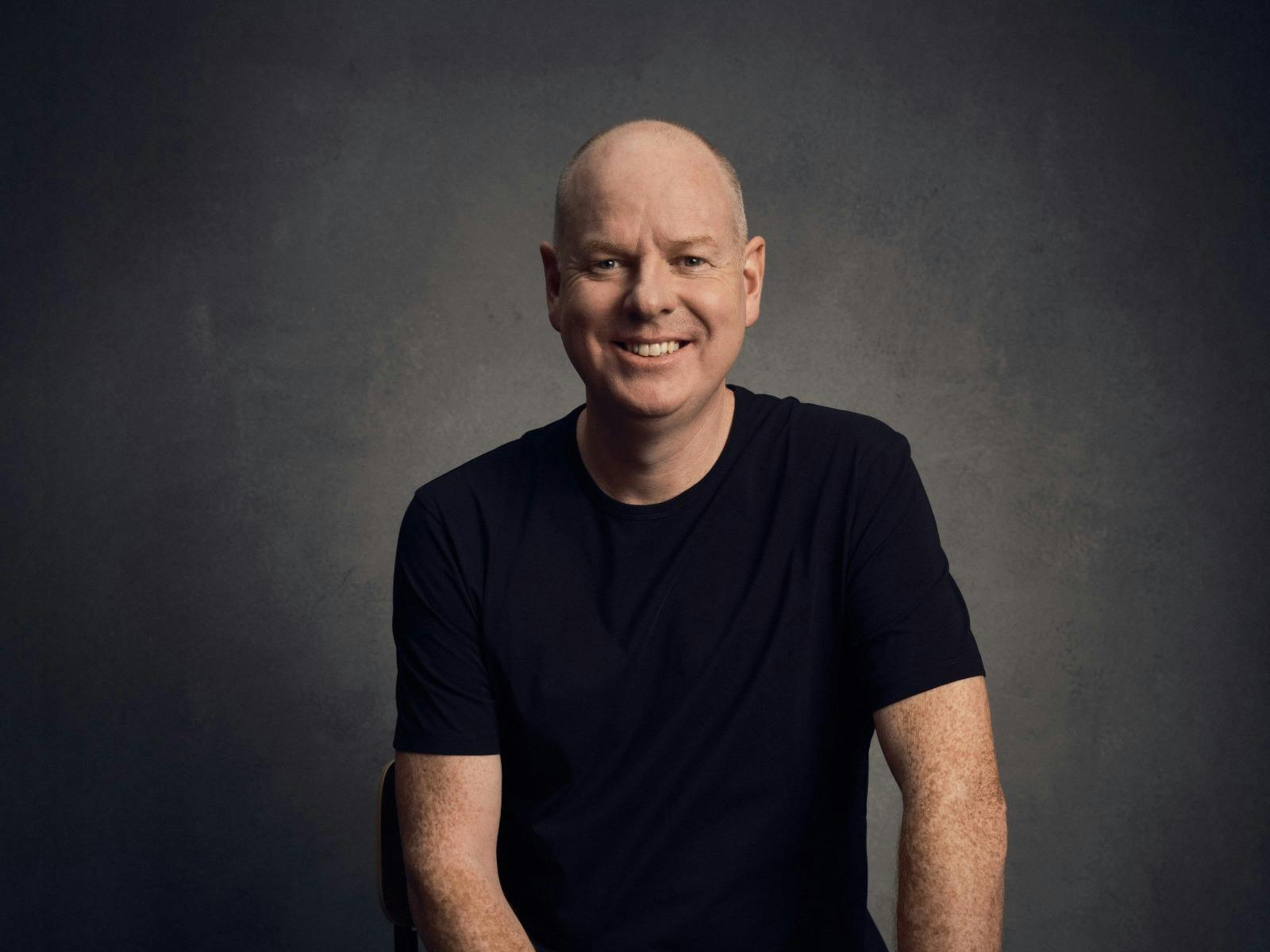 Comedian Tom Gleeson sitting on a stool, smiling Comedian Tom Gleeson sitting on a stool, smiling