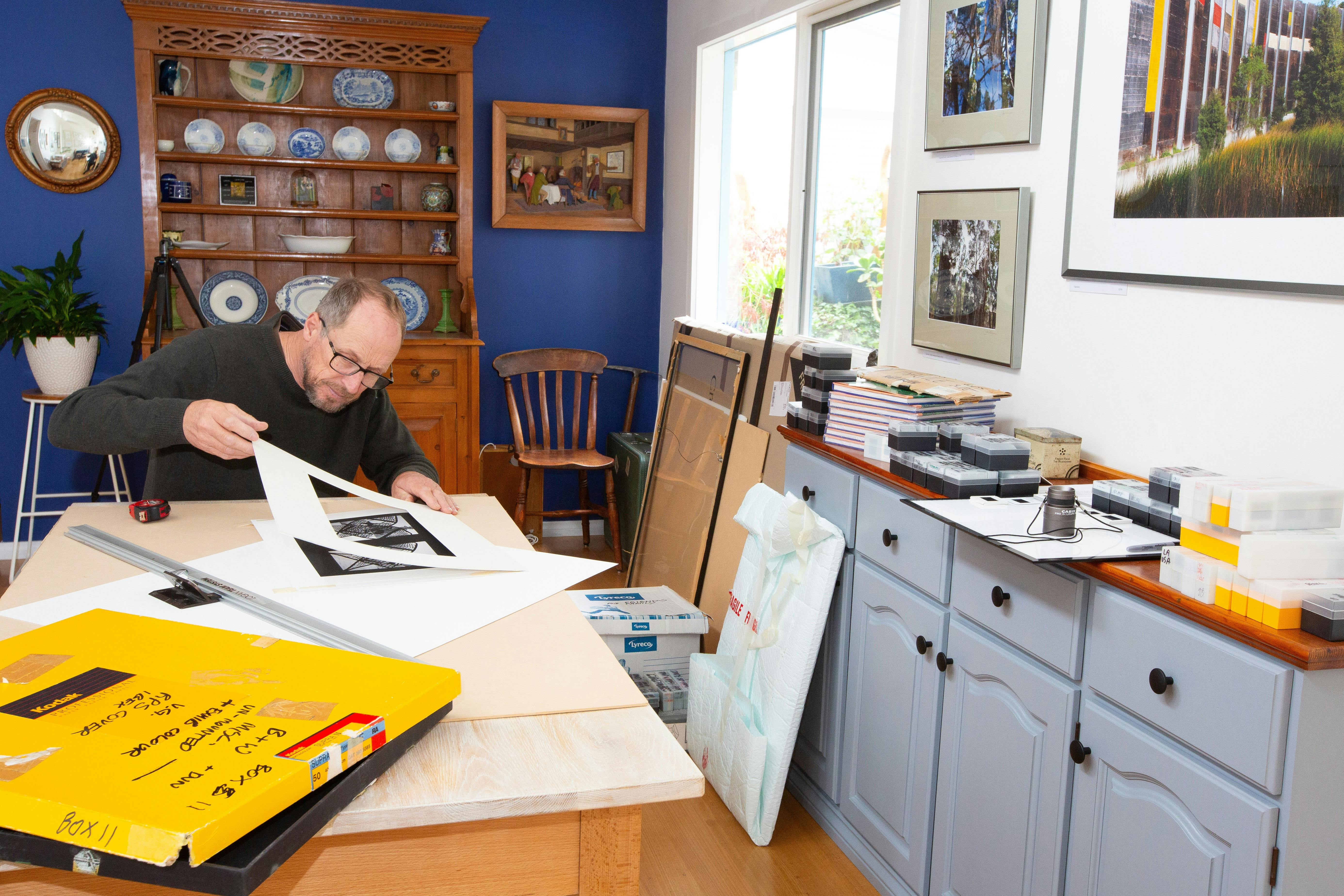 Photographer examines a black-and-white print at his studio table, surrounded by archives and framed