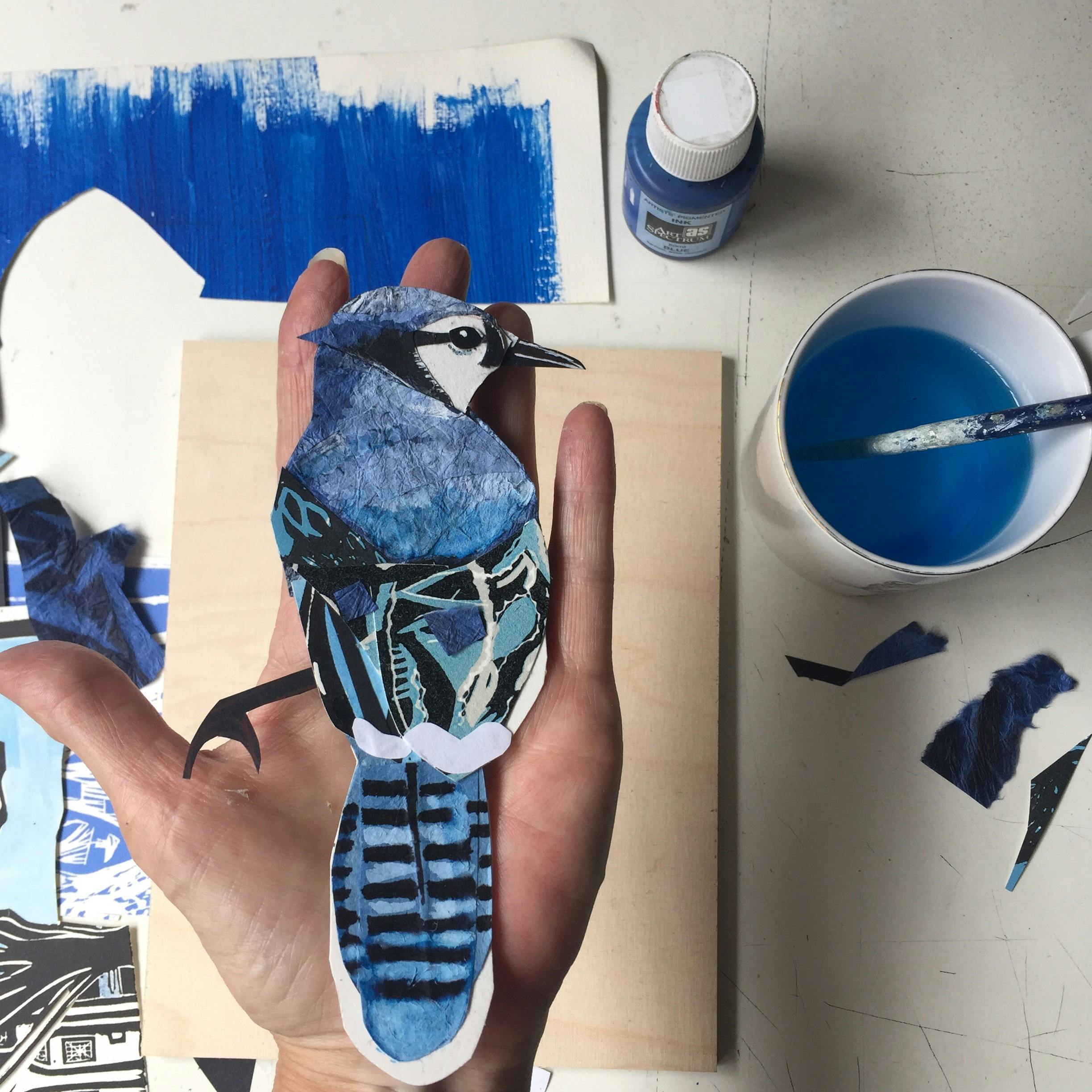 A hand holds a blue painted paper bird with a cup of blue ink with a brush to the right