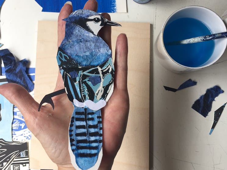 A hand holds a blue painted paper bird with a cup of blue ink with a brush to the right