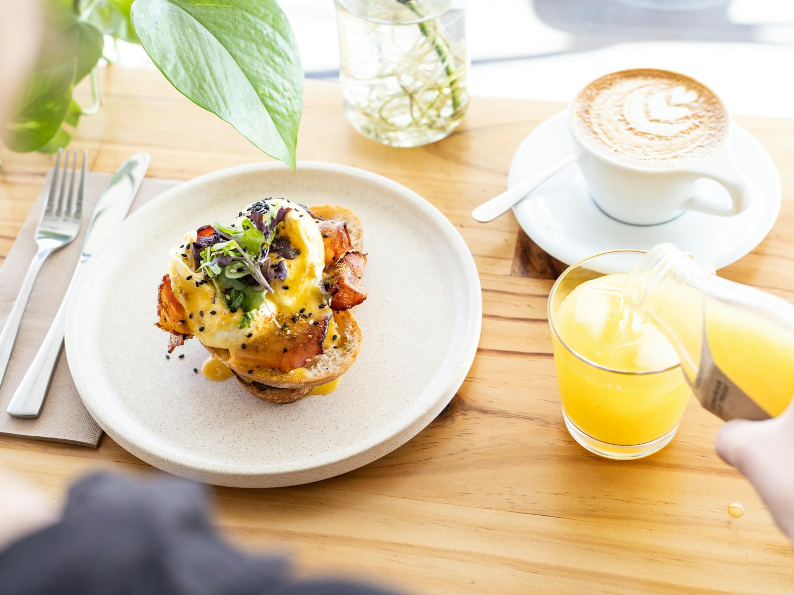 An eggs benedict served on a cream plate with a flat white and a glass of cold-pressed orange juice