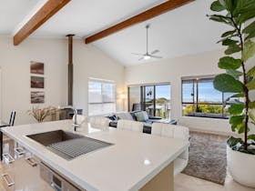Bright kitchen and living area with island bench, ceiling fan, timber beams, dining table, sofa