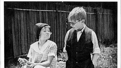 Black and white image of girl smirking and sitting down and boy with glasses looking down at her