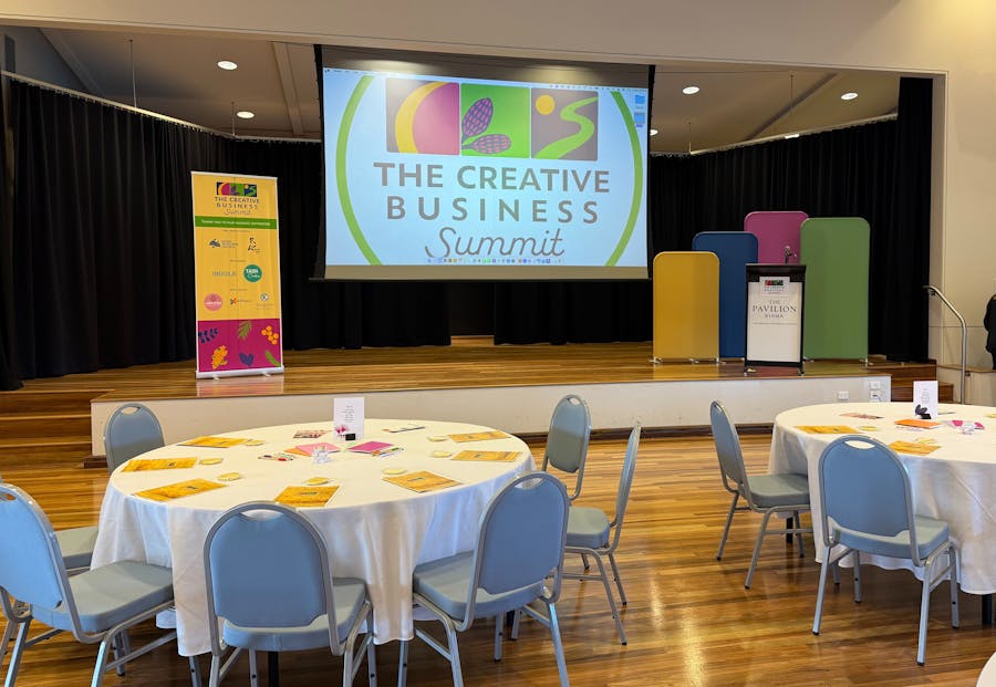 A stage with projector screen, conference tables & chairs. Bright set design in a light, airy room