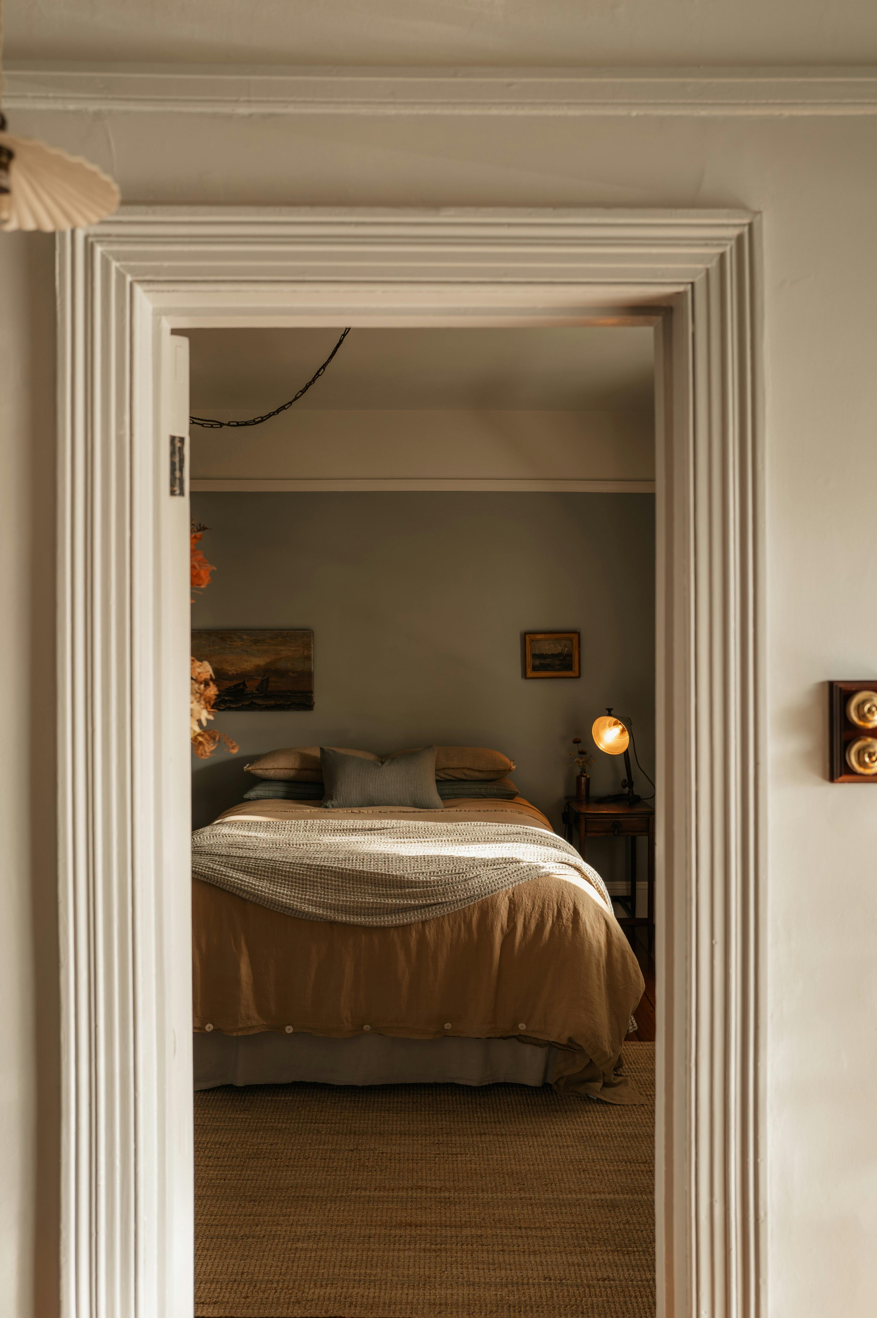Bedroom viewed through doorway with bed, bedside lamp and neutral tones.