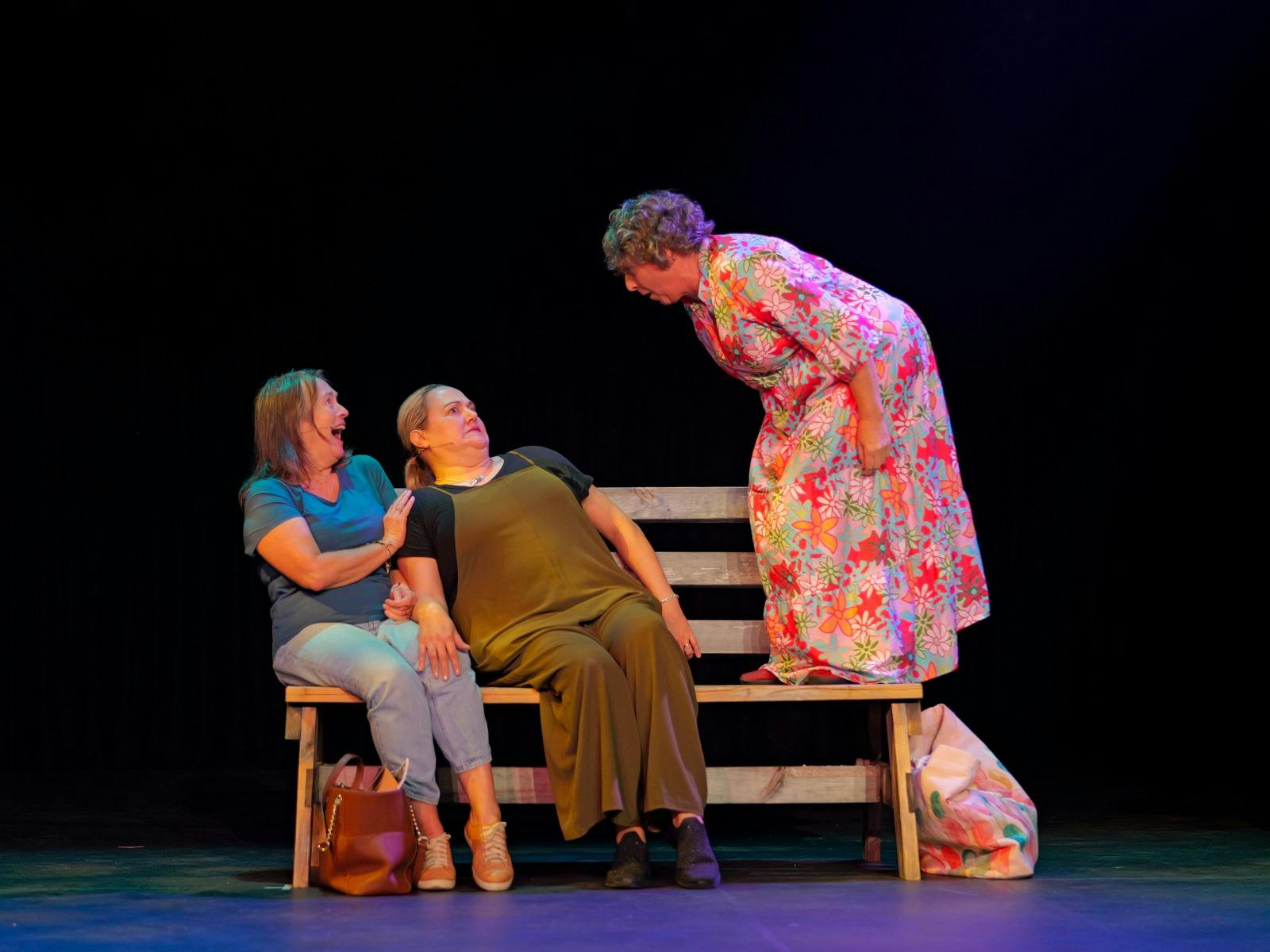 两名女子坐在长椅上，身体向后倾斜，背对着站在长椅上的第三名女子。