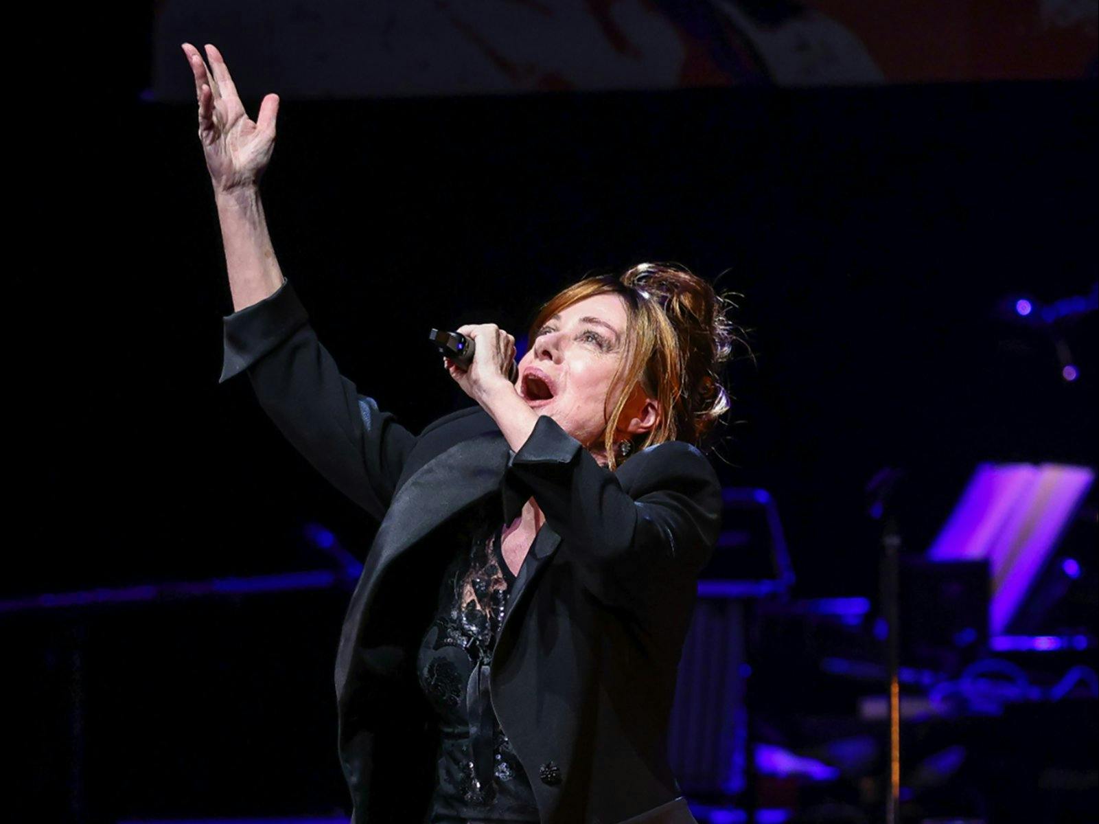 Performer singing into a microphone, one hand in the air, looking above. Purple music stand in back.