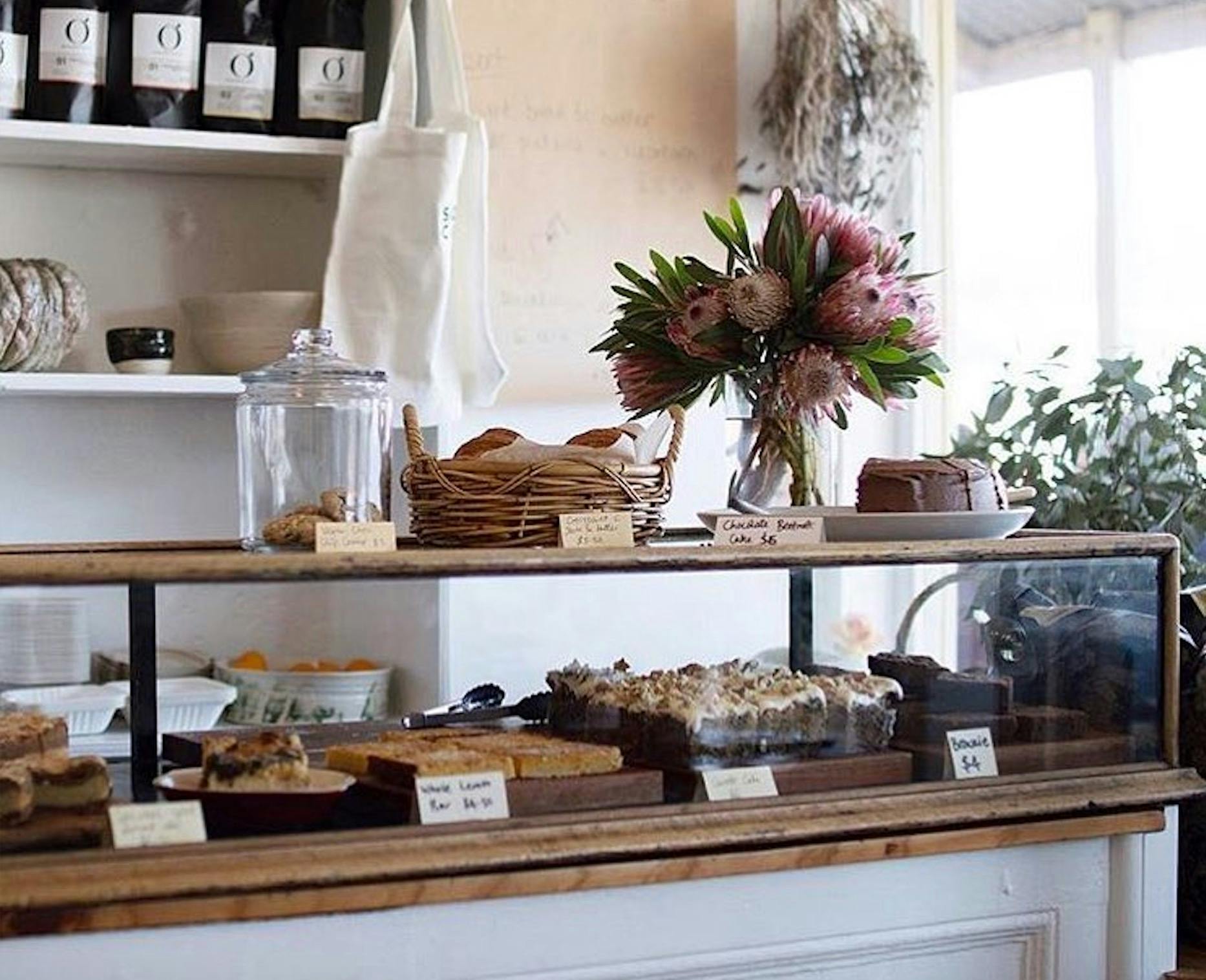 Cake cabinet full of homemade cakes. Fresh flowers sit on the bench.