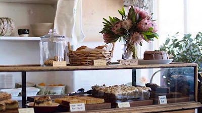 Cake cabinet full of homemade cakes. Fresh flowers sit on the bench.
