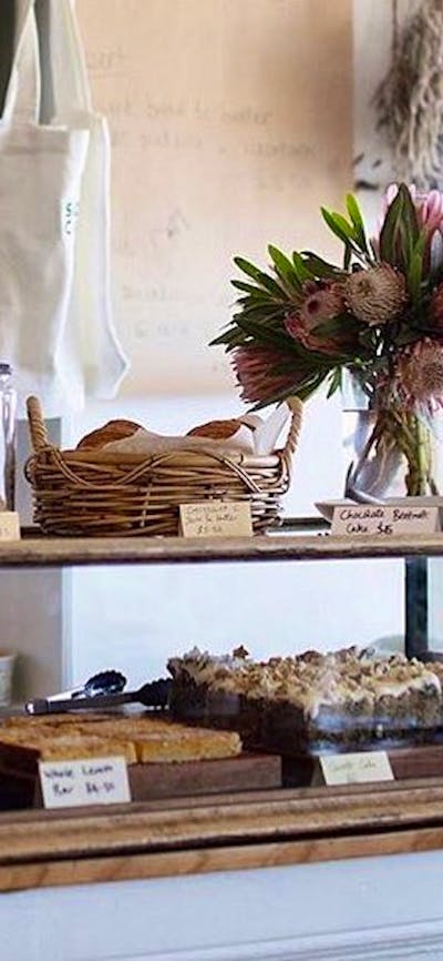 Cake cabinet full of homemade cakes. Fresh flowers sit on the bench.
