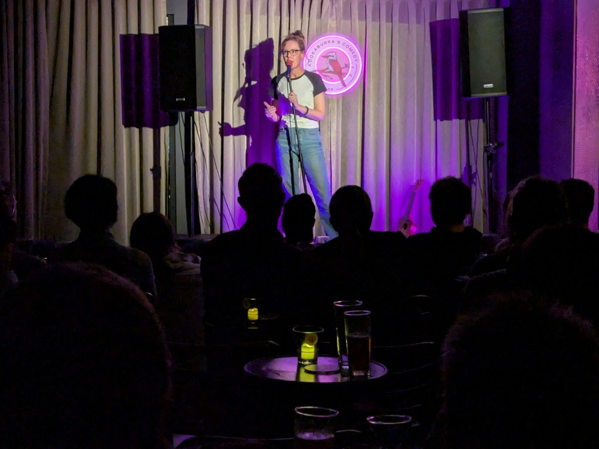 A comedian at the microphone by a glowing kookaburra sign, with the audience in silhouette