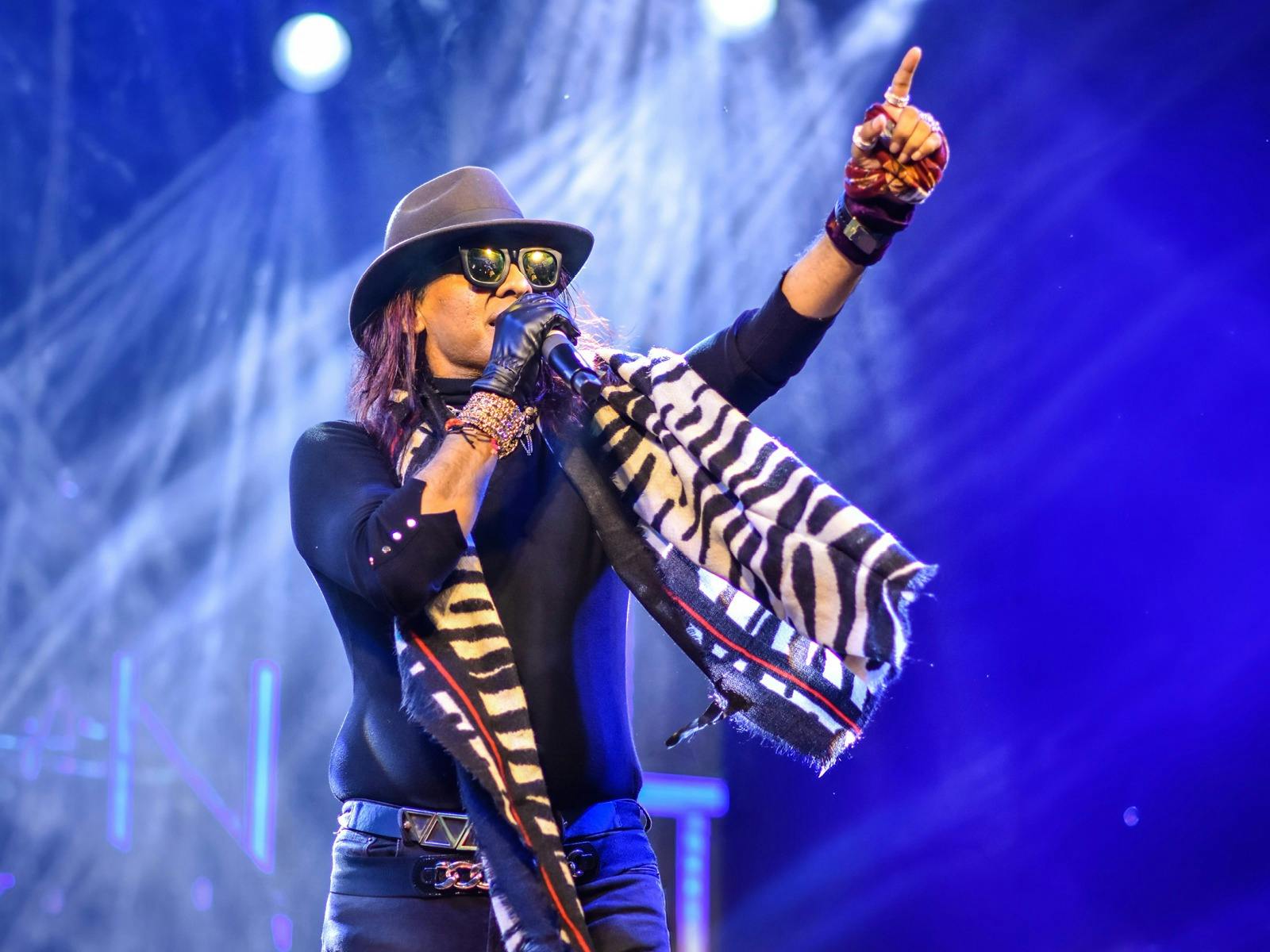 A south east Asian man wearing black jeans, shirt and hat and a zebra striped scarf