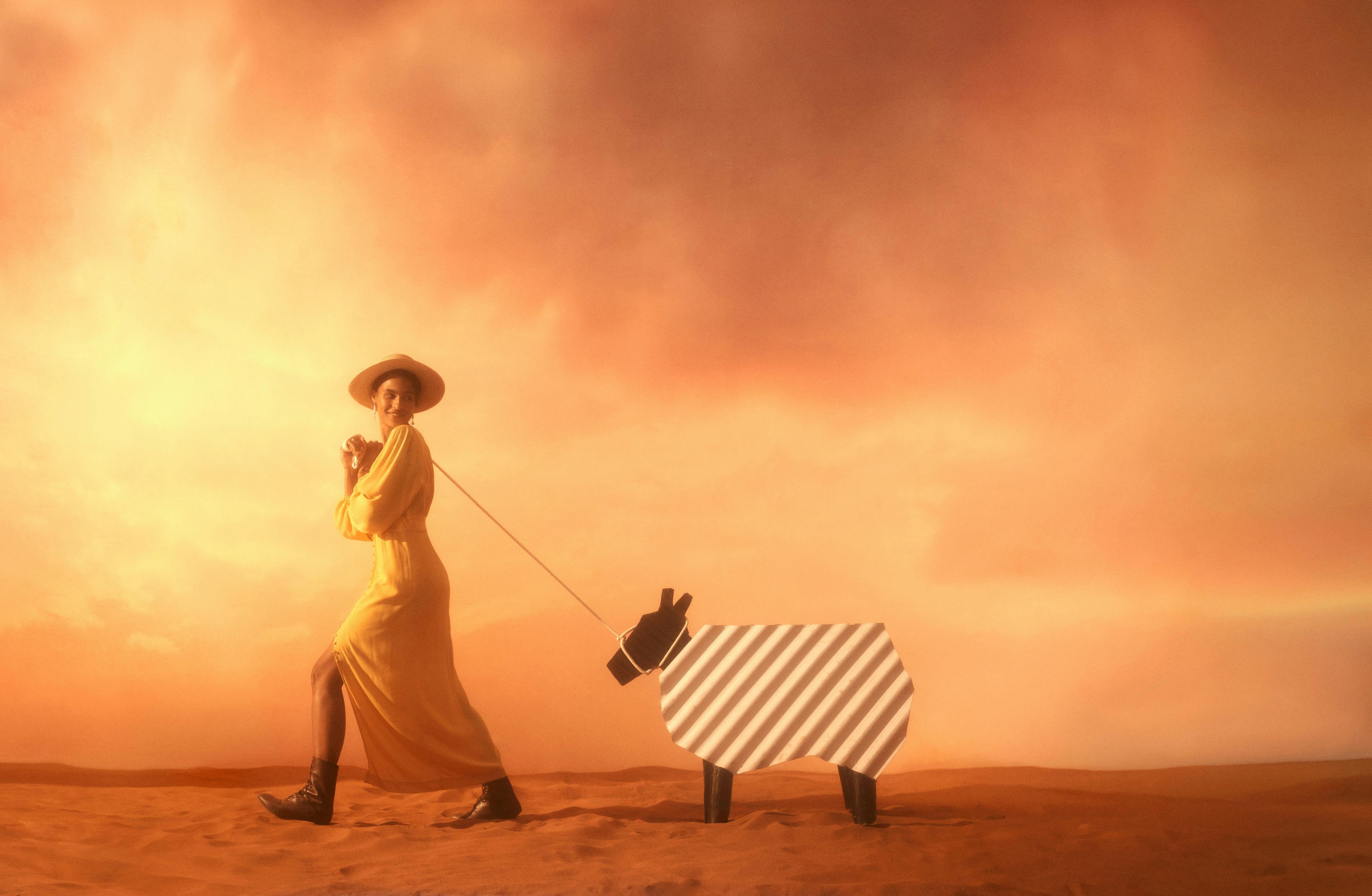 A woman walks across the sand at sunset leading a stylised, corrugated sheep