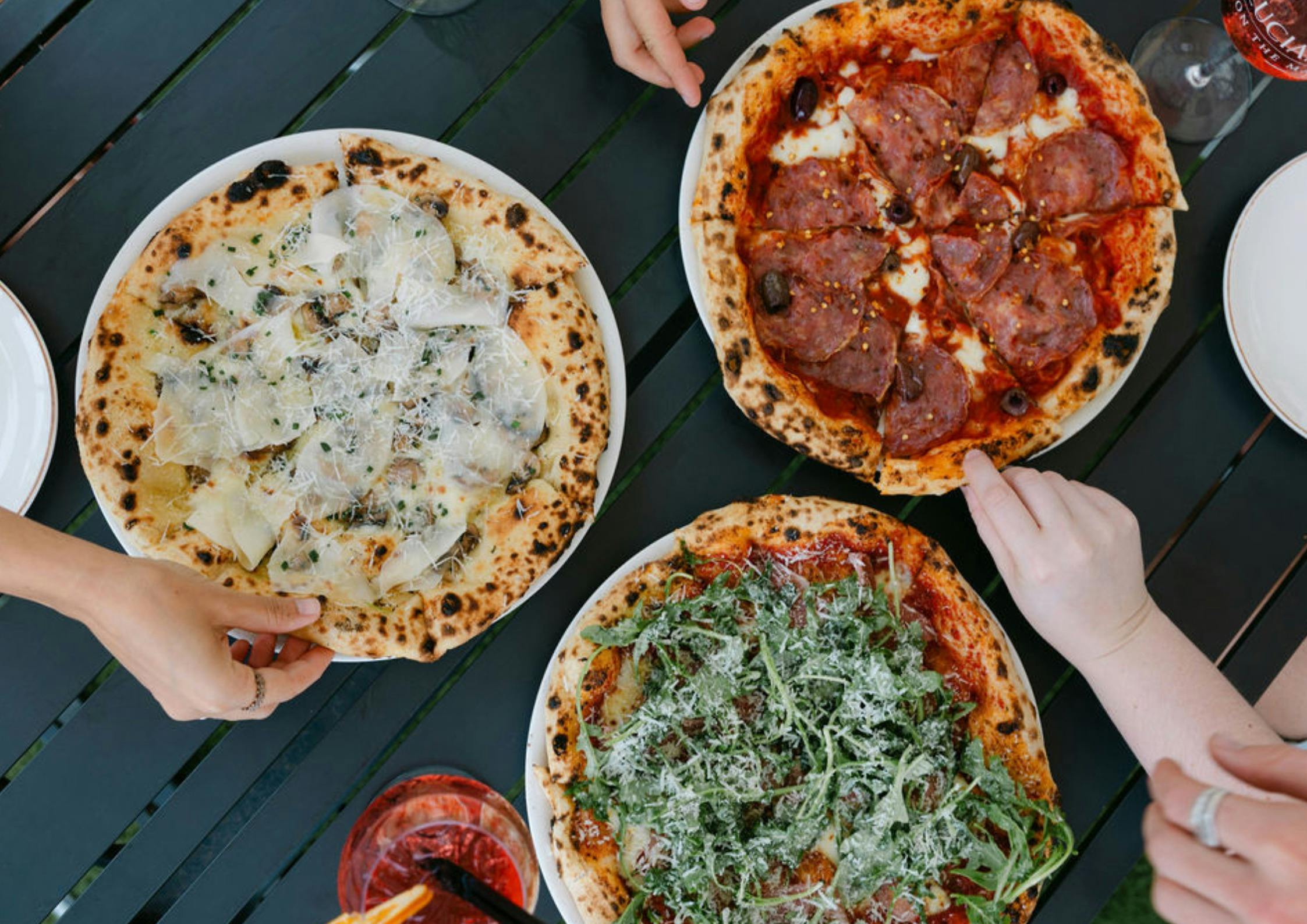 hands reaching for pizza slices of 3 different pizzas