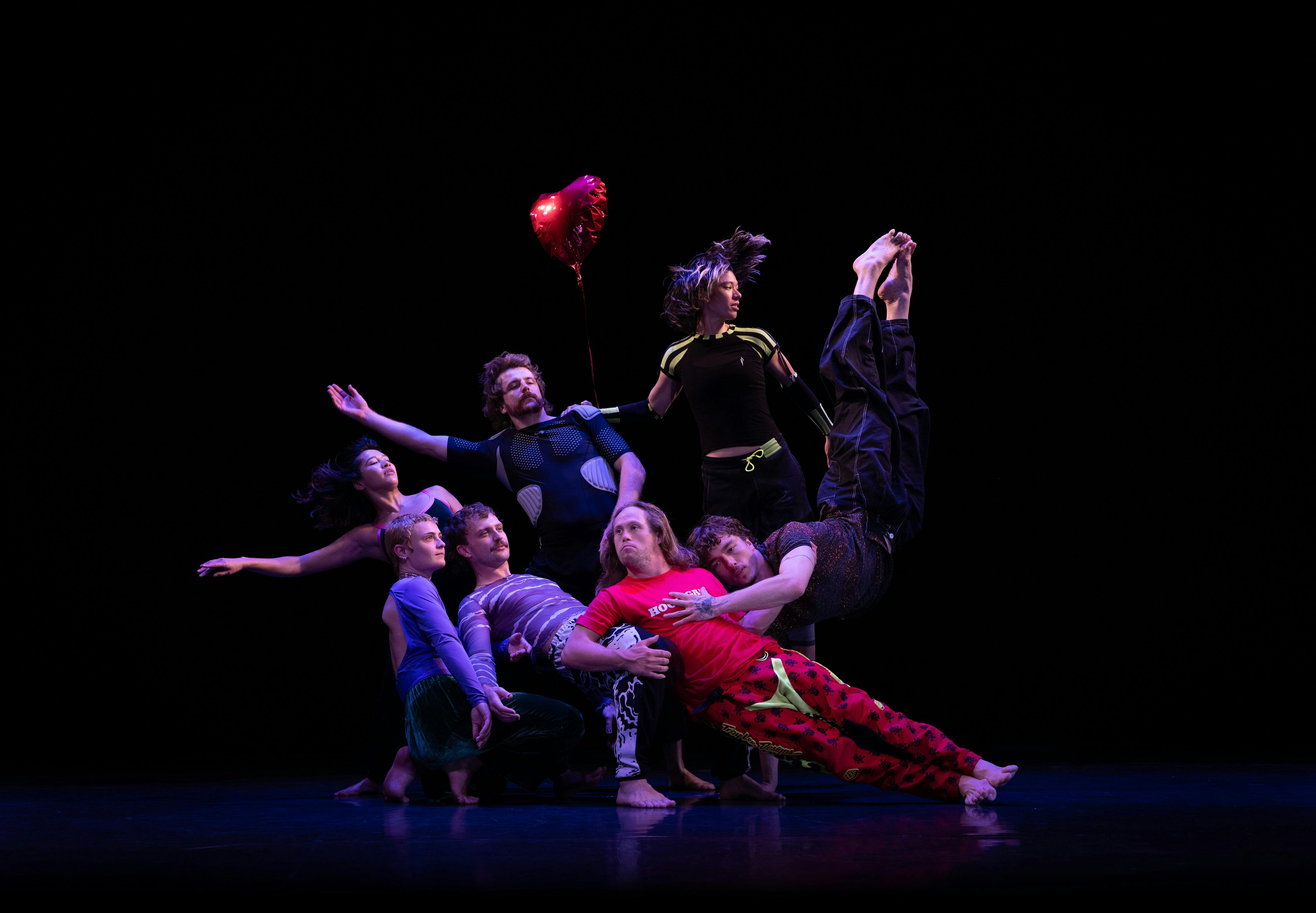 A group of dancers pose close together on stage. Some levitate. One holds a red heart balloon