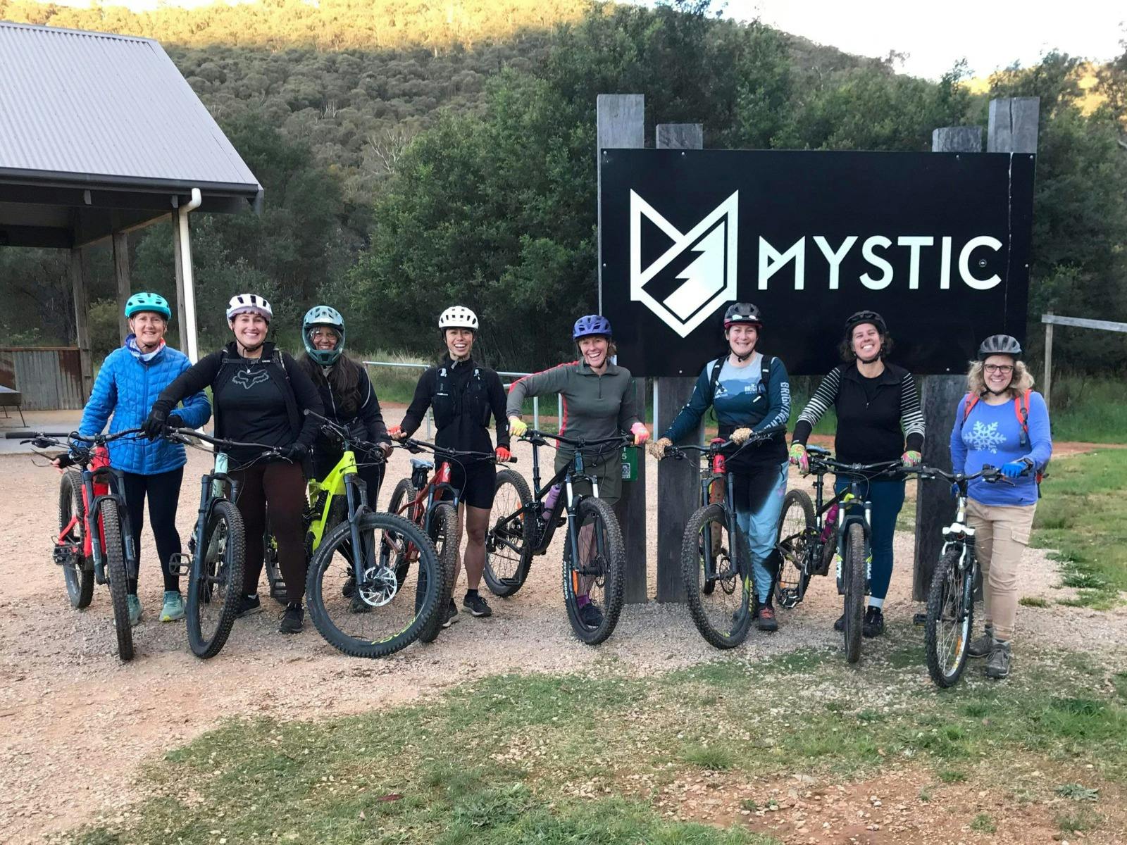 A group of women with bikes stand in front of a black sign that reads MYSTIC.