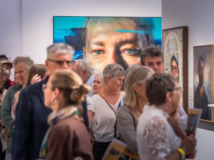 A group of people look at paintings while a portrait of a large face looks over them.