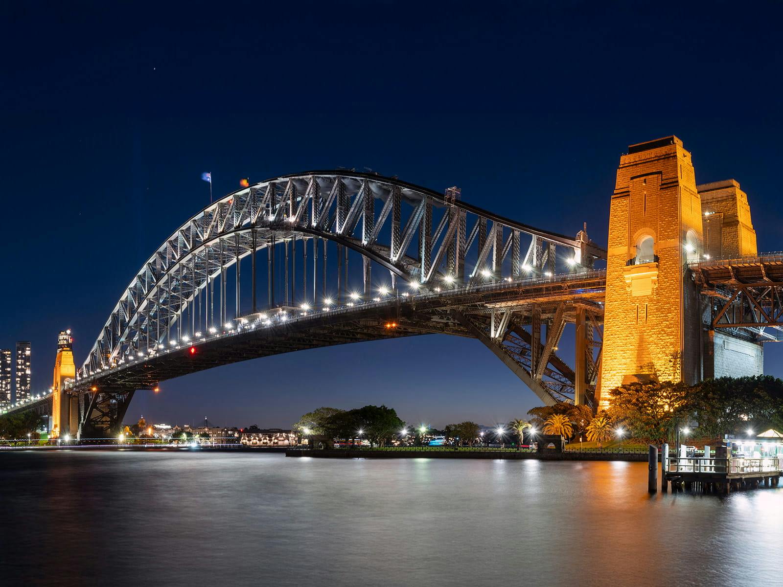 Sydney Harbour Bridge illuminated at night with reflections on calm harbour water.