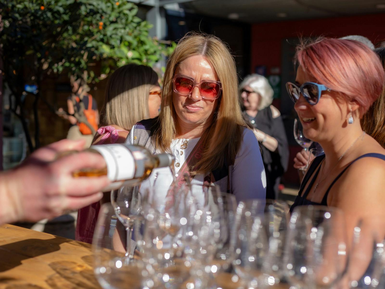 At an outdoor wine tasting, a woman pours wine as two women in sunglasses watch and smile