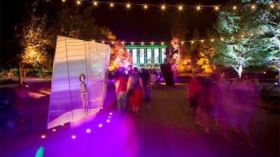 People walk past sculpture bathed in purple light. National Library of Australia in the background