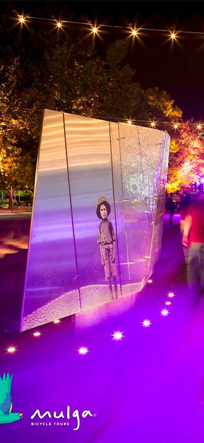 People walk past sculpture bathed in purple light. National Library of Australia in the background