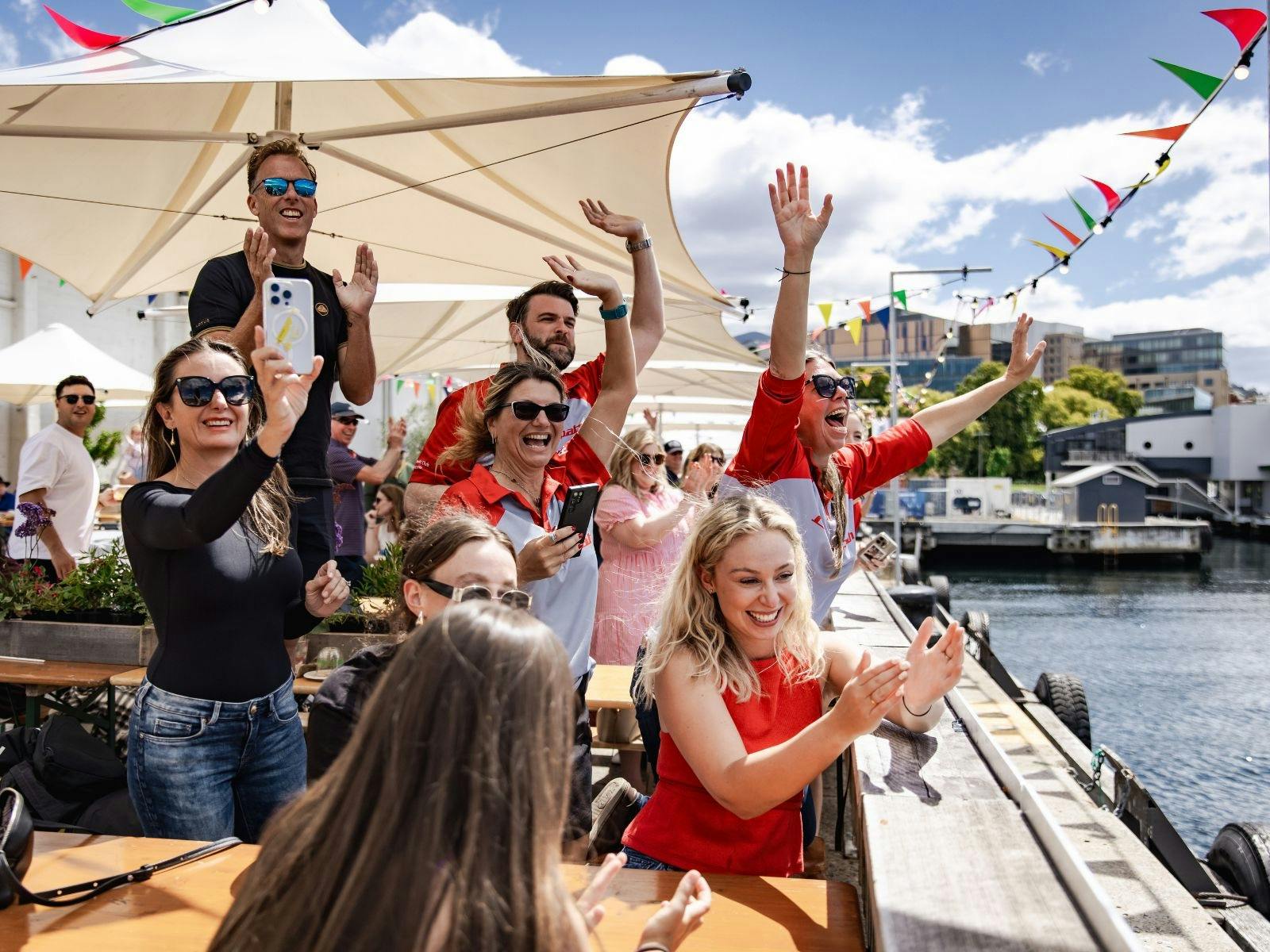 Smiling crowds wave from Hobart’s pier, facing the water during a lively festival moment.