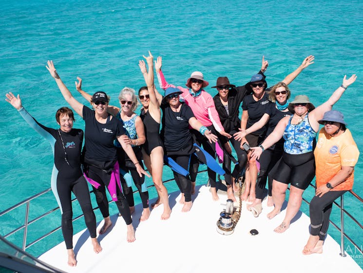 Women on the bow of 3 Islands Whale Shark Boat, hands in the air, wearing wet suits, blue water