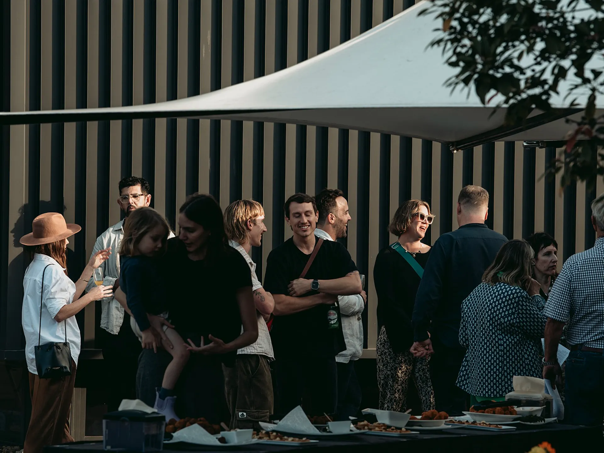 A crowd of people stand under a white umbrella talking outside a minimalist dark grey building.