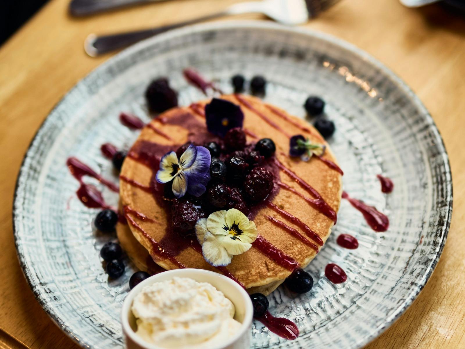 Birds eye view of pancakes with flowers, berry coulee drizzled on top, ramekin of cream on the side