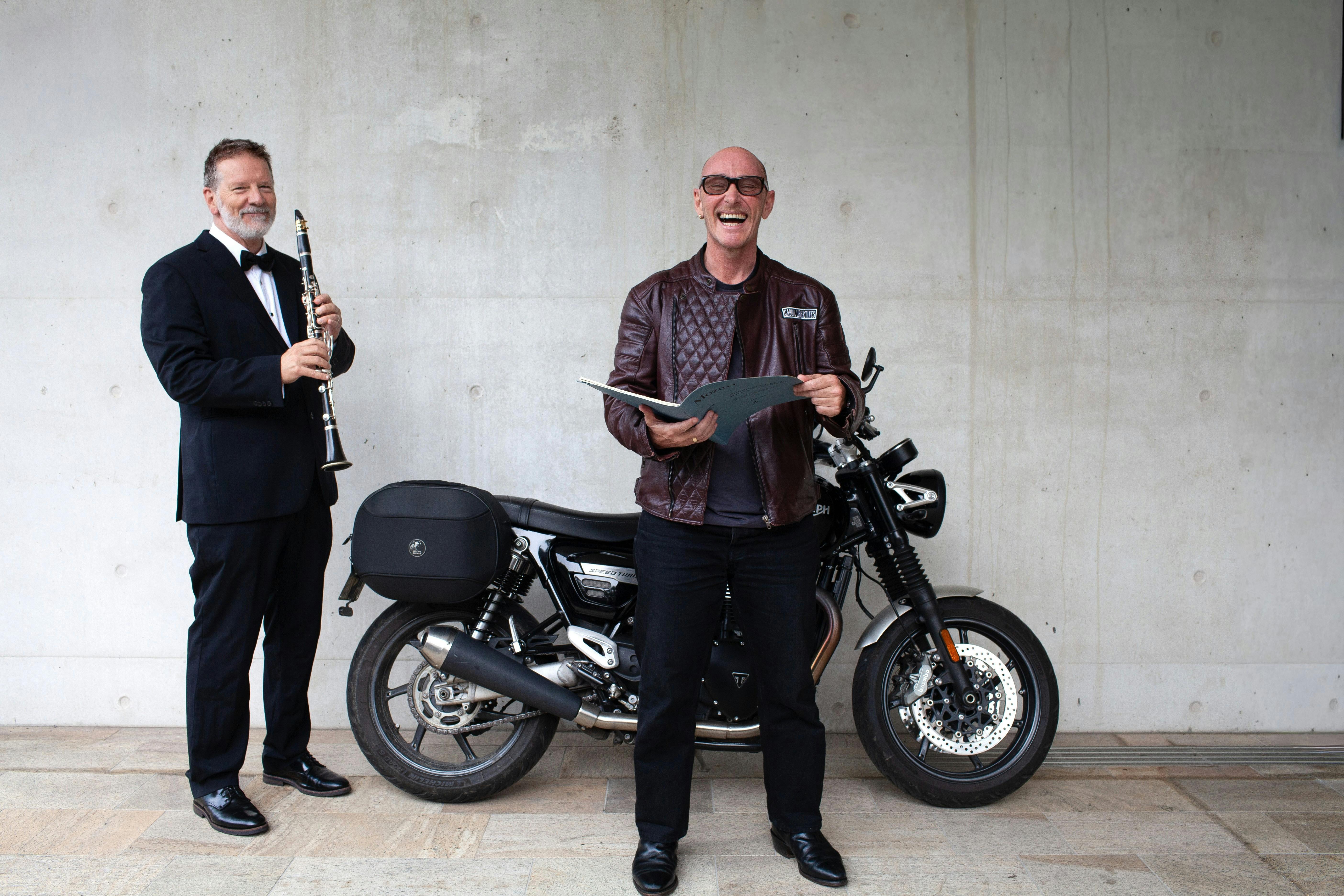Two men stand next to a motorcycle, one in a tuxedo with a clarinet and another in a leather jacket