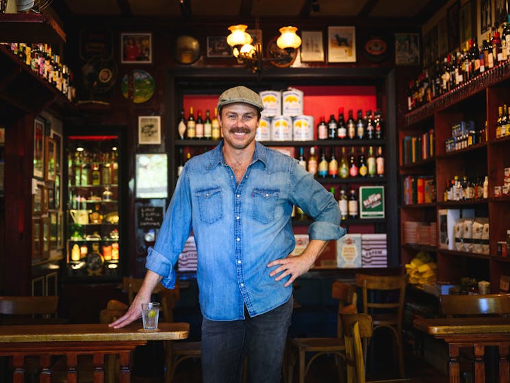 Smiling man standing inside bar with bottles, blue shirt and cap
