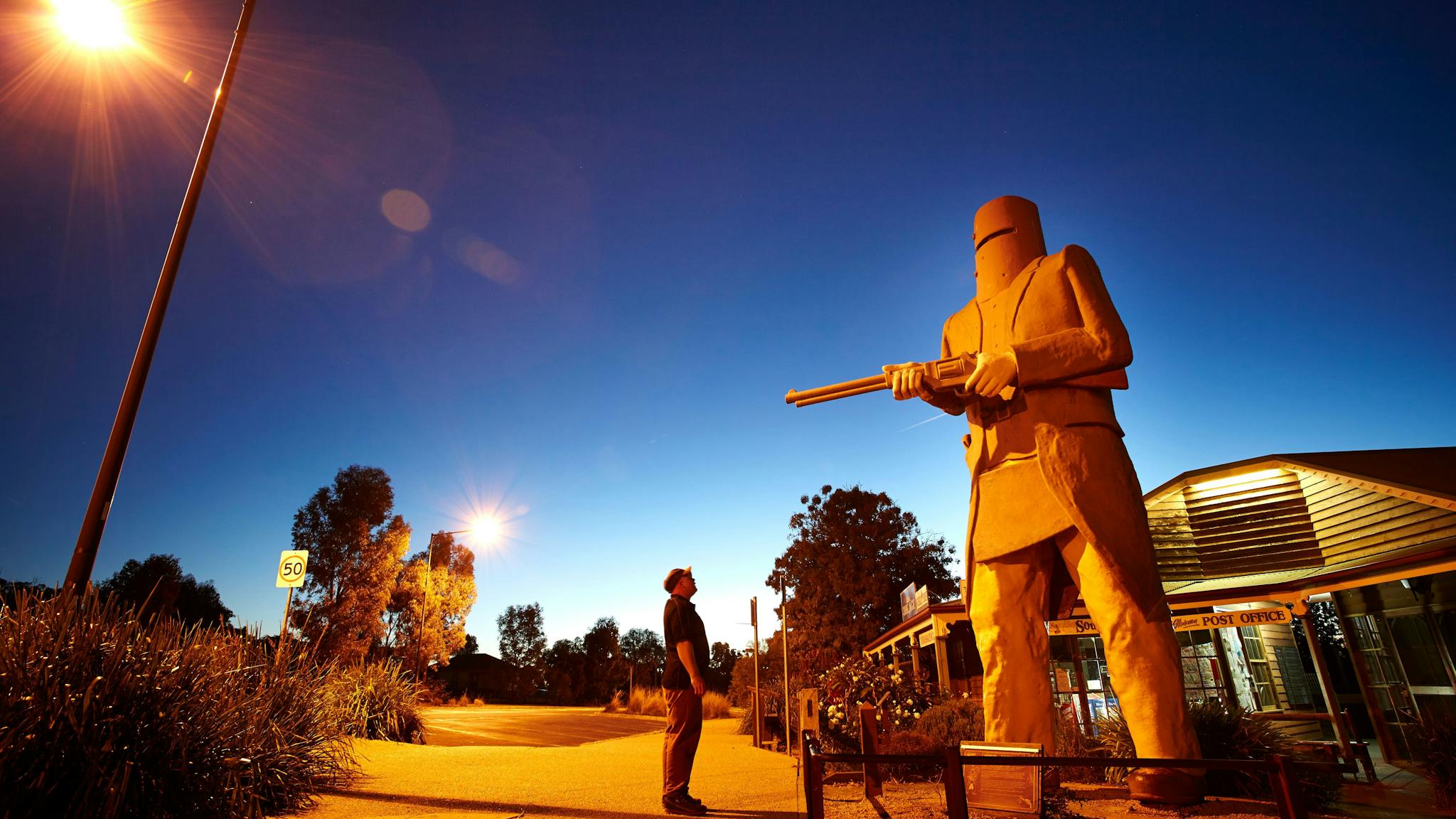 Person standing looking up at huge Ned Kelly Statue, shrouded by street lights as it gets dark.